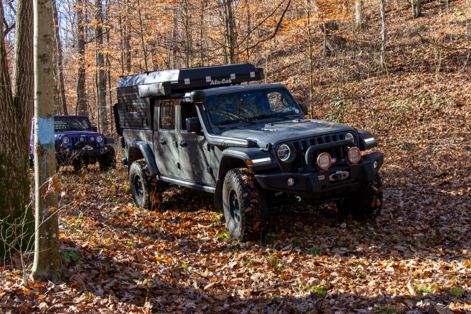 Lifted Jeep Gladiator with an Alu-Cab camper shell parked on a leaf-covered trail in the autumn woods, another Jeep visible in the background